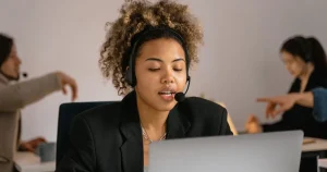 Atendente com headset falando com cliente em frente ao notebook, em escritório com outros atendentes ao fundo