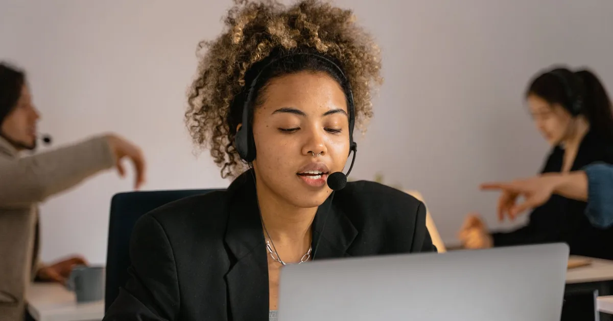 Atendente com headset falando com cliente em frente ao notebook, em escritório com outros atendentes ao fundo