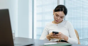 Profissional respondendo clientes pelo celular no escritório, com o notebook aberto ao lado