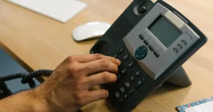 Mão discando em um telefone IP sobre a mesa do escritório, ao lado do teclado